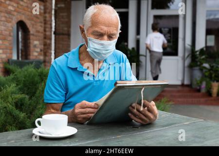 Älterer Mann in Schutzmaske mit einer Tasse Kaffee oder Tee in der Hand sitzt am Tisch des Straßencafés und liest die Speisekarte Stockfoto