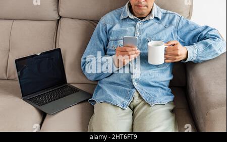 Aufnahme eines Geschäftsmanns, der ein Mobiltelefon benutzt, während er sich auf dem Sofa entspannt und Kaffee zu Hause getrunken hat Stockfoto