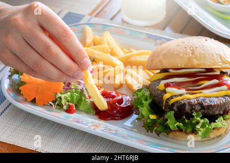 Eine Frauenhand taucht Pommes in Ketchup, und es gibt einen großen und schönen Hamburger mit einem Cutlet und Salat auf dem Tisch. Ein appetitlich, aber schädlich Stockfoto