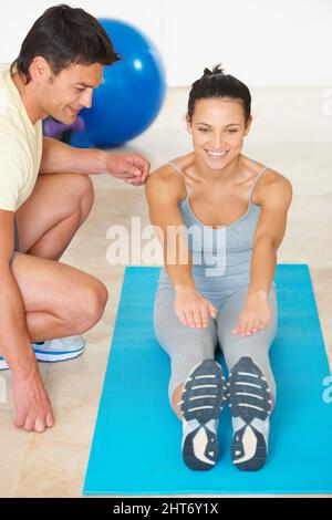 Geschmeidig und fit werden. Ein Fitnesstrainer, der einer jungen Frau zeigt, wie sie eine Übung macht. Stockfoto