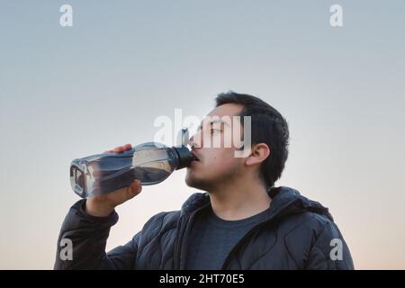 Aufnahme eines Mannes in schwarzer Jacke, der Wasser aus einer Plastikflasche trinkt Stockfoto