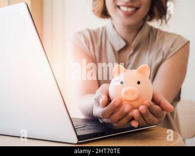 Rosa Sparschwein hält und gibt von asiatischen Frauen die Hände mit einem glücklichen Lächeln, während sie einen Laptop-Computer auf einem Holzschreibtisch benutzen. Investitionstechnologie, savi Stockfoto