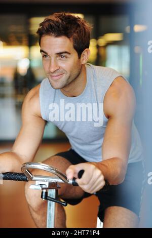 Er fühlt sich großartig in seiner Ausbildung. Ein lächelnder, hübscher junger Mann, der Sportkleidung trägt und im Fitnessstudio ein Heimtrainer benutzt. Stockfoto