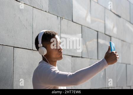 Junger hispanischer Mann mit Kopfhörern, der mit seinem Smartphone ein Selfie auf der Straße machte Stockfoto