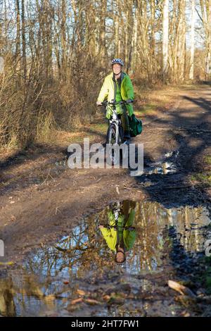 Frau über fünfzig macht mit ihrem E-Bike eine Radtour auf einem schlammigen Weg und durch Pfützen, Lüneburg, Niedersachsen, Deutschland Stockfoto