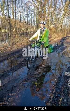 Frau über fünfzig macht mit ihrem E-Bike eine Radtour auf einem schlammigen Weg und durch Pfützen, Lüneburg, Niedersachsen, Deutschland Stockfoto