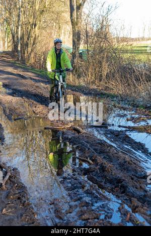 Frau über fünfzig macht mit ihrem E-Bike eine Radtour auf einem schlammigen Weg und durch Pfützen, Lüneburg, Niedersachsen, Deutschland Stockfoto