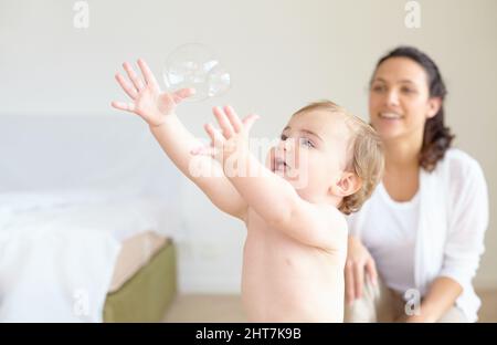 Was für Wunder die Welt für sie bereit hält. Ein entzückendes Mädchen, das nach einer Blase greift und ihre Mutter im Hintergrund lächelt - Copyspace. Stockfoto