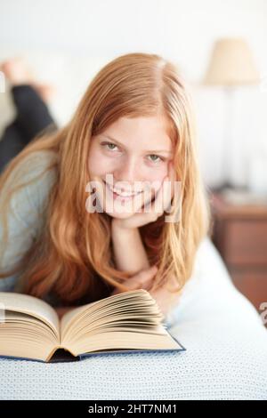 Genießen Sie die Welt der Fiktion. Porträt eines jungen Rotschopf, der ihr Buch liest und auf ihrem Bett liegt. Stockfoto