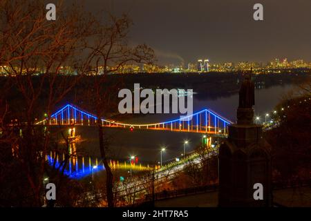9.. April 2019. Kiew, Ukraine. Die Parkovyi-Brücke über den Dnjepr erleuchtete die blauen und gelben Farben der ukrainischen Flagge. Im Vordergrund ist die Silhouette des Großen Denkmals Wolodymyr zu sehen, im Hintergrund die Stadt Ukraine. Stockfoto
