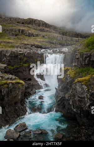 Vertikale Aufnahme eines schäumenden Wasserfalls, der unter dem düsteren Himmel über die schwarzen, moosigen Felsen gleitet Stockfoto