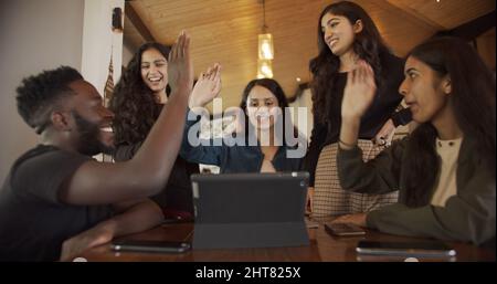 Eine Gruppe junger südasiatischer Menschen, die hohe Fünfen machen und in einem Café mit einem Tablet auf dem Tisch sitzen Stockfoto