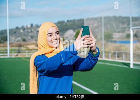 Glückliche ethnische Frau, die Selfie auf dem Smartphone auf dem Sportplatz macht Stockfoto