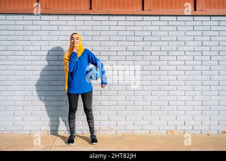 Lächelnde arabische Frau mit Fußballball in der Nähe des Backsteingebäudes Stockfoto