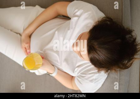 Draufsicht auf eine Schwangere, die mit einem Glas frischen Orangensaft in der Hand auf dem Sofa sitzt Stockfoto