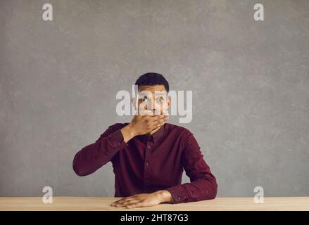 Junger afroamerikanischer Geschäftsmann sitzt im Büro und schaut schockiert auf die Kamera Stockfoto