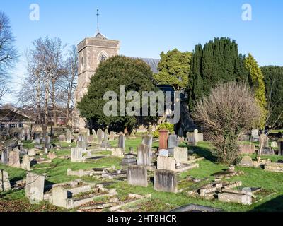 Die Sonne scheint auf der St. Saviour's Church und Eibenbäume und Grabsteine auf dem Kirchhof in Coalpit Heath in South Gloucestershire. Stockfoto