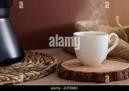 Nahaufnahme einer Tasse aus weißem Porzellan, die auf einem Stück Holz mit heißem, dampfendem Kaffee platziert wird. Daneben befindet sich eine schwarze Kaffeekocher und ein Sacktuch. Im Bac Stockfoto