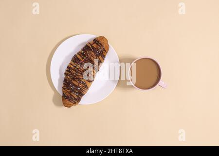 Schokoladencroissant auf dem Teller mit heißem Kaffee auf pastellbeigem Backgorund. Köstliches Snack- und Süßspeisen-Konzept Stockfoto