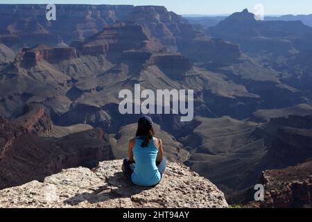 Ein Mädchen sitzt mit gekreuzten Beinen und blickt auf die Klippen, Buttes und Felsschichten hinunter in die Tiefen des Grand Canyon am Shoshone Point auf dem Sou Stockfoto