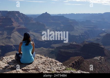 Ein Mädchen sitzt mit gekreuzten Beinen und blickt auf die Klippen, Buttes und Felsschichten hinunter in die Tiefen des Grand Canyon am Shoshone Point auf dem Sou Stockfoto