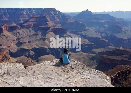 Ein Mädchen sitzt mit gekreuzten Beinen und blickt auf die Klippen, Buttes und Felsschichten hinunter in die Tiefen des Grand Canyon am Shoshone Point auf dem Sou Stockfoto