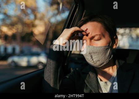 Geschäftsfrau mit starken Kopfschmerzen Schmerzsymptomen und schützender Gesichtsmaske auf dem Rücksitz eines Autos, selektiver Fokus Stockfoto