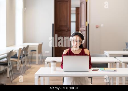Serious Asian Student Mädchen in Kopfhörern beobachten pädagogische via Internet auf Laptop in der Bibliothek Stockfoto