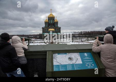 Blick auf die Hauptkathedrale der russischen Streitkräfte (Kathedrale der Auferstehung Christi) im Patriot Park, Oblast Moskau, Russland. Touristen und Kinder in Winterjacken stehen auf einer Aussichtsplattform und blicken mit stationären Ferngläsern auf die monumentale dunkelgrüne Kathedrale mit goldenen Kuppeln unter bewölktem Himmel Stockfoto