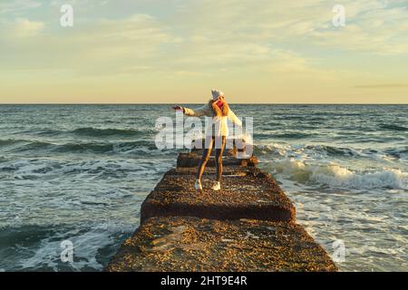 Eine Frau mit blondem Haar in einem Hut und einem Pelzmantel geht an einem Steinbrecher entlang des Meeres Stockfoto