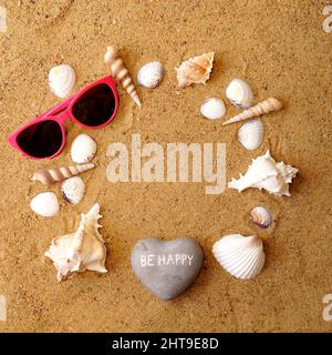 Ein Stein in Form eines Herzens mit der Inschrift sei glücklich, Gläser und verschiedene Arten von Muscheln liegt auf dem Strandsand in Form eines Kreises. In Stockfoto