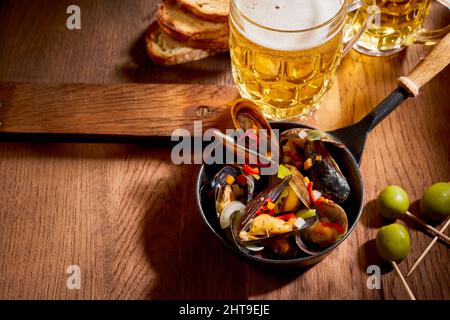Schüssel mit leckeren Muscheln serviert auf Holztisch mit einem Glas Lagerbier und grünen Oliven Stockfoto