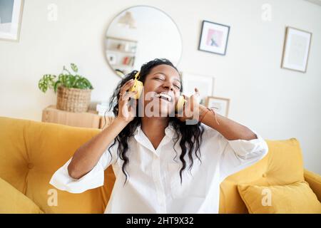 Fröhliche junge Frau, die mit Kopfhörern Musik hört und tagsüber zu Hause auf der weichen Couch tanzt. Stockfoto