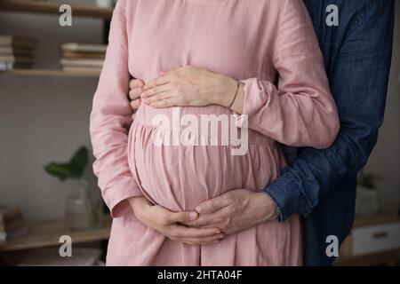 Ehemann umarmt Schwangere in rosa Leinenkleid von hinten Stockfoto