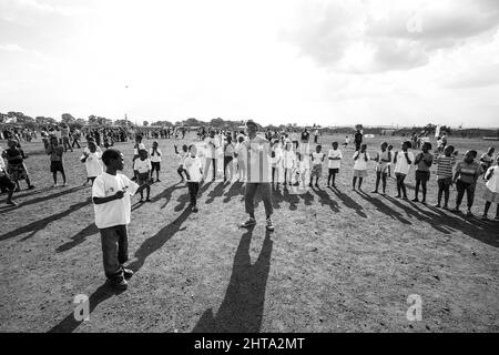 Graustufenaufnahme eines freiwilligen Fußballtrainers, der mit jungen afrikanischen Kindern auf dem Schulhof arbeitet Stockfoto