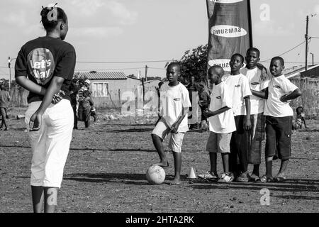 Graustufenaufnahme eines freiwilligen Fußballtrainers, der mit jungen afrikanischen Kindern auf dem Schulhof arbeitet Stockfoto