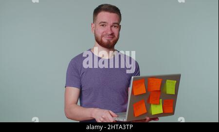 Erschöpfter Mann Freiberufler mit eingeklebten Aufkleber Notizen mit Laptop-Computer mit Konzentration Problem kreative Krise. Nervöser Erwachsener Kerl verwirrt durch b Stockfoto