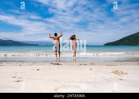 Lateinisches Paar, das auf den weißen Sandstrand und das blaue Meer, die Insel Anchieta in Brasilien, zuläuft. brasil ilha Stockfoto