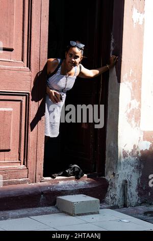 Eine junge Frau hält ihr Handy und lehnt sich an die Tür, während ihr Hund auf dem Boden in der Tür ihres Hauses in Havanna, Kuba, liegt. Stockfoto