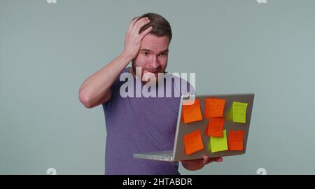 Erschöpfter Mann Freiberufler mit eingeklebten Aufkleber Notizen mit Laptop-Computer mit Konzentration Problem kreative Krise. Nervöser Erwachsener Kerl verwirrt durch b Stockfoto