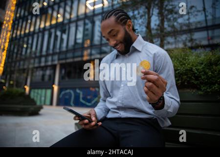 Ein Kryptowährungshändler ist mit den Ergebnissen seiner Investitionen zufrieden, er hat sein Smartphone, eine Bitcoin- und ethereum-Münze in der Hand Stockfoto