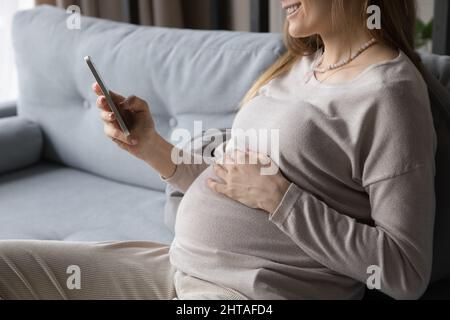 Glückliche junge, erwartete Mutter, die ihr Handy benutzt und sich auf dem bequemen Sofa entspannt Stockfoto