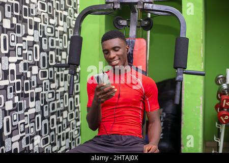 Der junge Afrikaner benutzt sein Telefon, während er in der Pause auf einem pec-Deck in einem Fitnessstudio sitzt Stockfoto