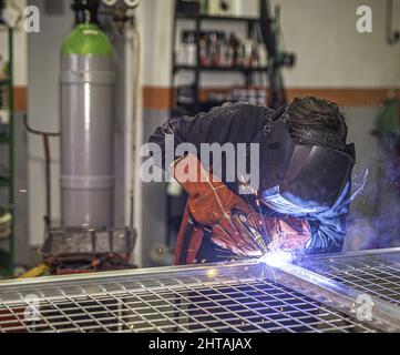 Professioneller Schweißer mit Gesichtsschutzmaske, der eine Tür in der Werkstatt schweißen kann. Stockfoto