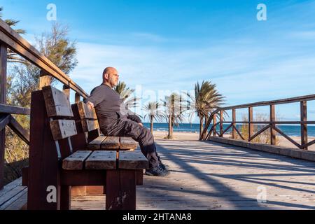 Kaukasischer Mann mittleren Alters, der auf einer Bank am Strand sitzt Stockfoto