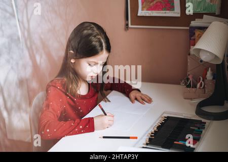 Hübsches Schulmädchen mit langen Haaren in rotem Kleid zieht am Tisch im Kinderzimmer zu Hause Stockfoto