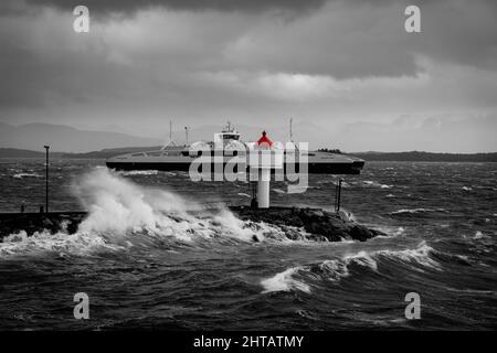 Eine Graustufenaufnahme eines Leuchtturms und eines Schiffes, das auf rauer See vorbeifährt Stockfoto