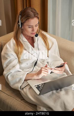 Eine Frau, die mit einem Smartphone in einem Headset mit einem Laptop arbeitet Stockfoto