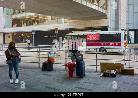 Hongkong, China. 27.. Februar 2022. Haushelfer schauen vor einem Polizeiauto im Central District in Hongkong auf ihre Telefone. Hongkong verschärft die Regeln der sozialen Distanzierung, um der schlimmsten Epidemie entgegenzuwirken, indem es unter allen Umständen Masken im Freien trägt und sich auf zwei Personen beschränkt. Kredit: SOPA Images Limited/Alamy Live Nachrichten Stockfoto