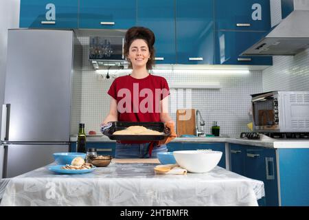 Porträt einer schönen, glücklichen jungen Frau, die ein Backblech mit Pizzateig in der Hand hält. Ansicht von unten. Das Konzept der Hausmannskost und Slow Food. Stockfoto